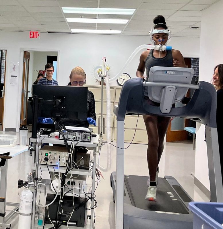 In a laboratory environment, a woman with tied-back blonde hair and glasses, wearing blue medical gloves, stands behind a computer. To her right, a young Black woman runs on a treadmill and wears a breathing apparatus. 