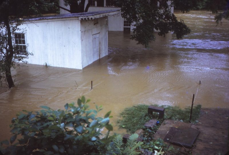 This photo shows a white shed and barn surrounded by murky, swirling brown flood waters.