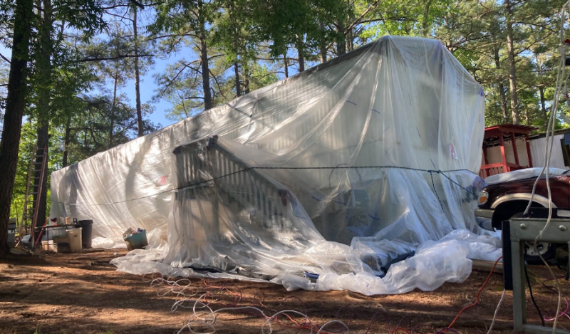 This photo shows a trailer home in a forested area completely draped in semi-transparent plastic. 