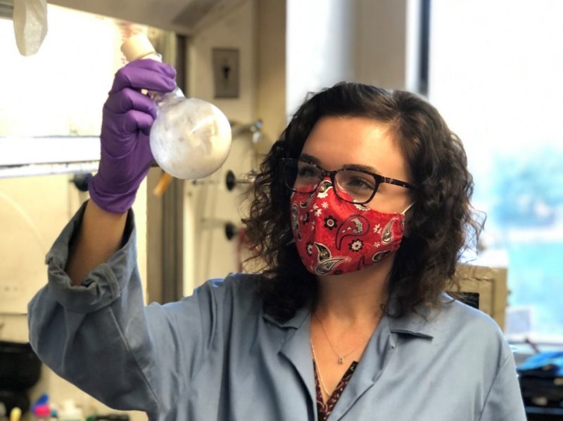 This photo shows a young white woman with shoulder length curly dark hair and dark-rimmed glasses. She is wearing a blue lab coat and a red paisley face mask and holding a flask filled with a white substance. 