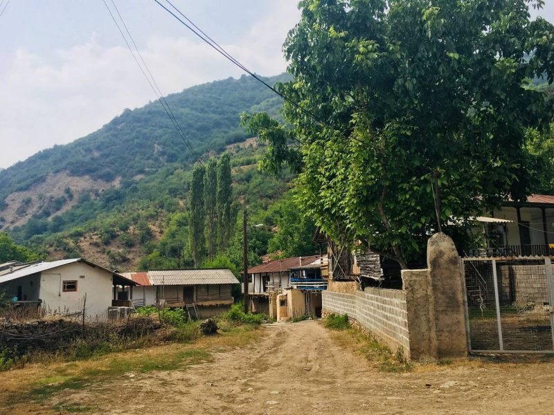 This photo shows a dirt road running between small buildings and a tree-covered mountain in the background.