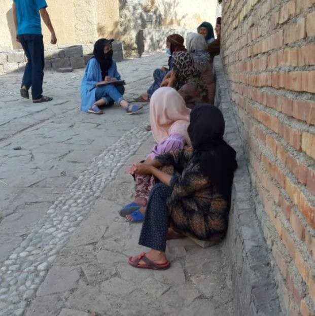 This photo shows six women wearing headscarves and seated on the ground with their backs to a brick wall. In front of them is another woman with a clipboard.