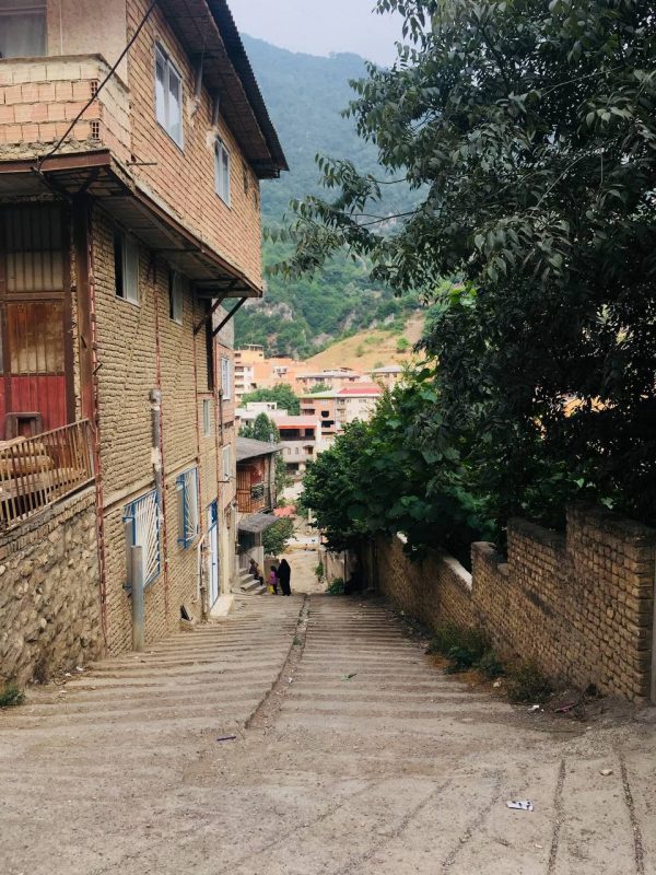 This photo shows a steep and narrow street between a building on the left and a wall on the right. More buildings can be seen below and a mountain in the background.