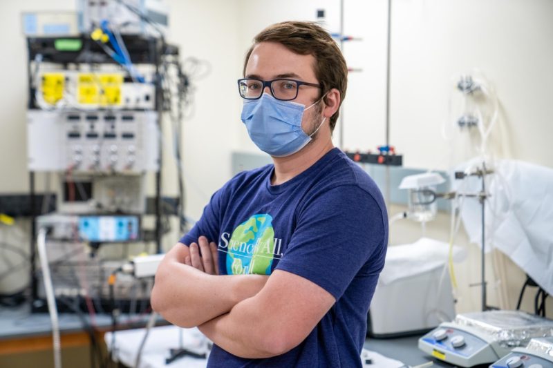 Ryan King, a young white male, wearing a blue shirt looks at the camera standing in his lab.