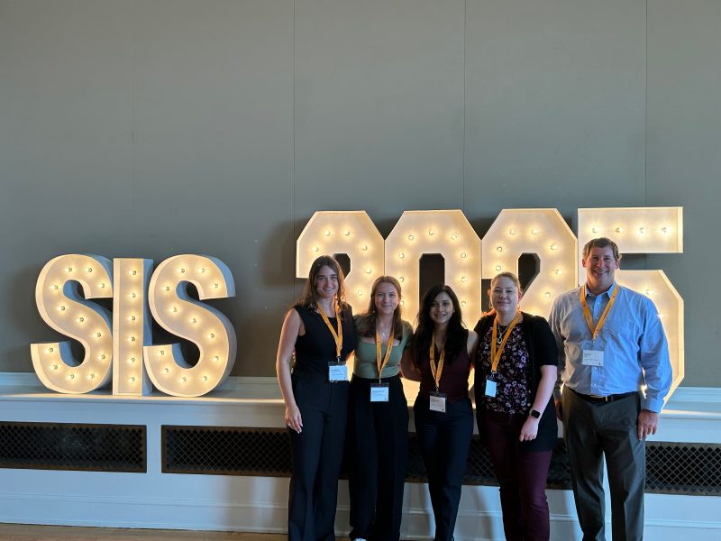 Image includes five smiling people in front of glowing letters spelling out SIS 2025 (Southeastern Immunology Symposium).