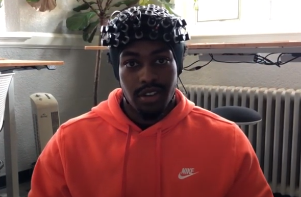 This photo shows a young Black male dressed in a bright orange Nike sweatshirt and wearing a bike-helmet-like contraption on his head. His head and shoulders are showing and he is looking directly into the camera.