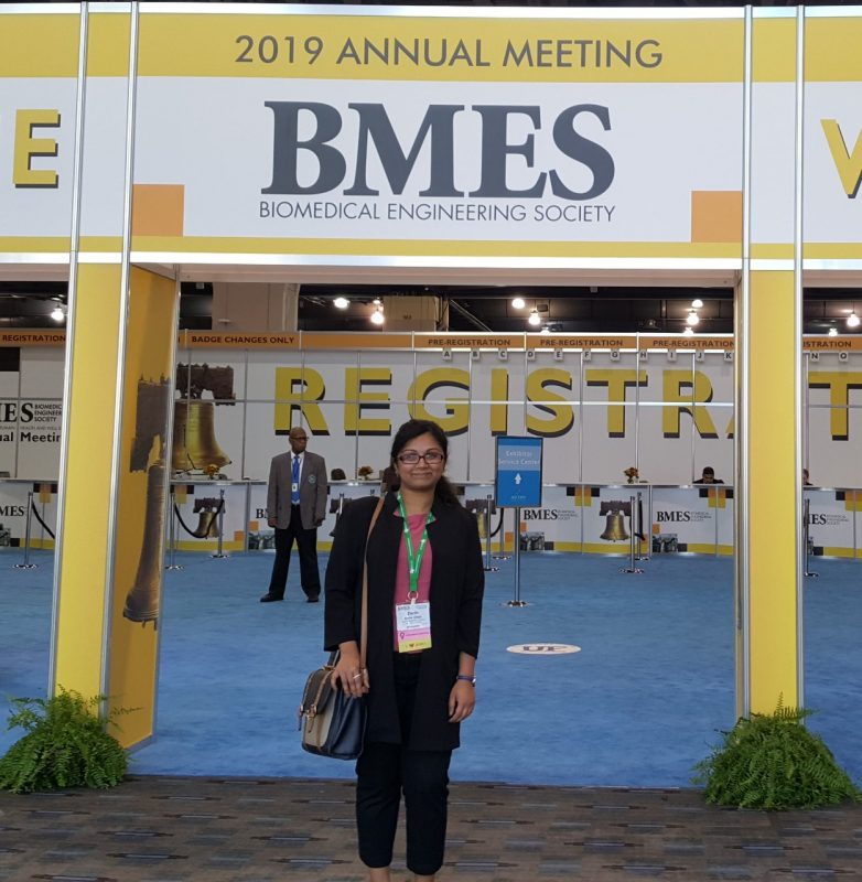 This photo shows a young woman dressed in black slacks, a black jacket, and a pink blouse standing under a sign that reads "2019 Annual Meeting BMES Miomedical Engineering Society." Behind her is a large room with a blue carpet and "Registration" on the wall. She has black hair, glasses, and medium brown skin. 