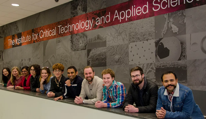 This photo shows 11 young adults from the waist up, leaning on a wall with their hands folded in front of them and smiling at the camera. Six appear to be women and five men, and a variety of nationalities and ethnicities/races are represented. Behind them on the wall is a banner that reads "The Institute for Critical Technology and Applied Science."