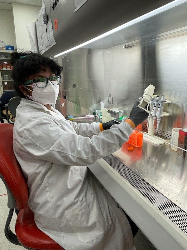 This photo shows a young woman wearing a white lab coat, white face mask, and gray gloves seated on a red office chair in front of a fume hood. She has black hair, glasses, and brown skin and is looking directly at the camera.