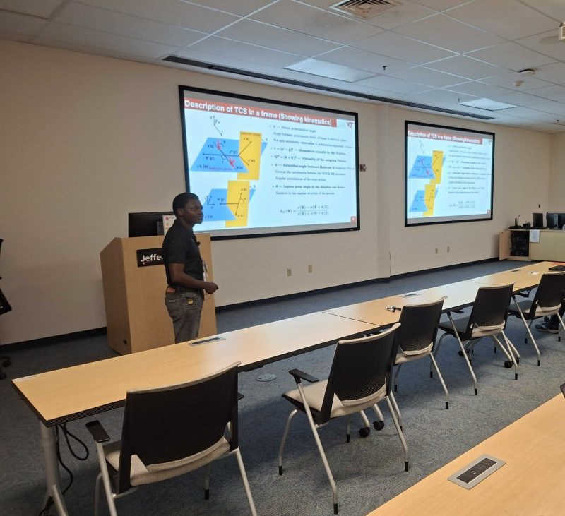Gyang, a physics researcher, stands in front of a classroom with two powerpoint slides titled "Description of TCS in a frame (showing kinematics)" projected behind him on the wall.