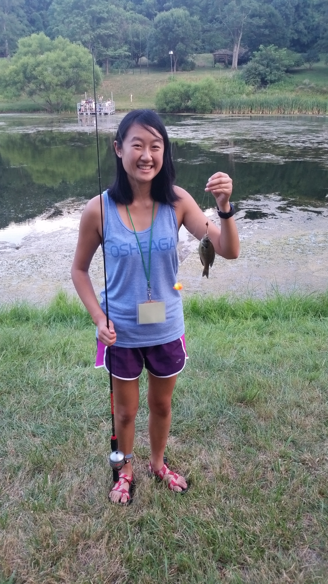 This photo shows a dark haired young woman holding a fishing pole in one hand and a fish on a line in the other. In the background is a pond. She is smiling. It appears to be summer.