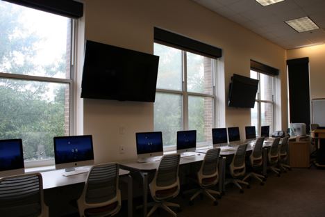 This studio shows a row of desks facing large windows on the edge of a room.  Eight chairs and eight computer monitors are visible, along with two large monitor screens on the wall in the space between the windows.