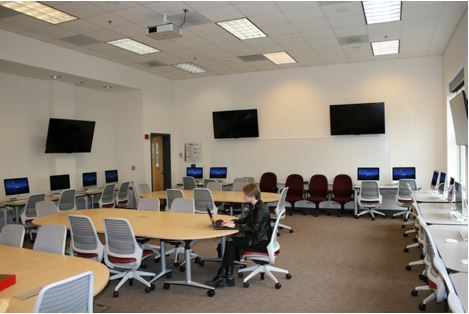 This photo shows a large classroom with desks and computer monitors lining the walls and three large tables, each with eight chairs, in the open space in the middle of the room.