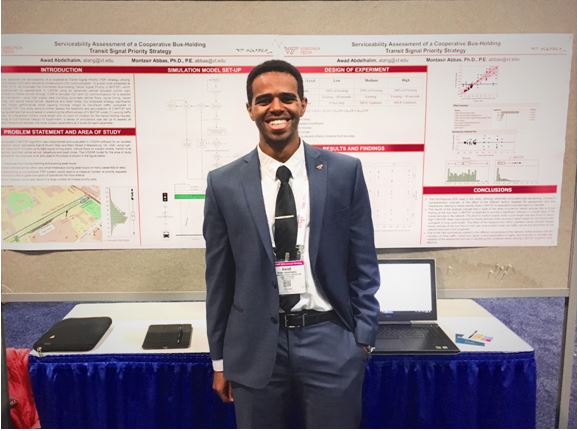 This photo shows a young man with brown skin and dark hair wearing a blue gray suit with a dark tie that has a conference name tag clipped to it. He is smiling  and is standing in front of a skirted conference-type table with research posters on the wall behind him.