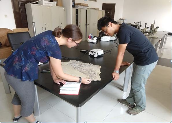 This photo shows a dark haired woman leaning over a fossil slab on a large black table. To the right in the photo is a black haired young man. The setting is a laboratory.