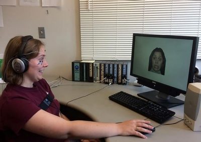 This photo shows a young white woman wearing earphones and a maroon t-shirt seated in front of a computer with an alarmed looking face on the screen. On the woman's face are electrodes with white wires attached.
