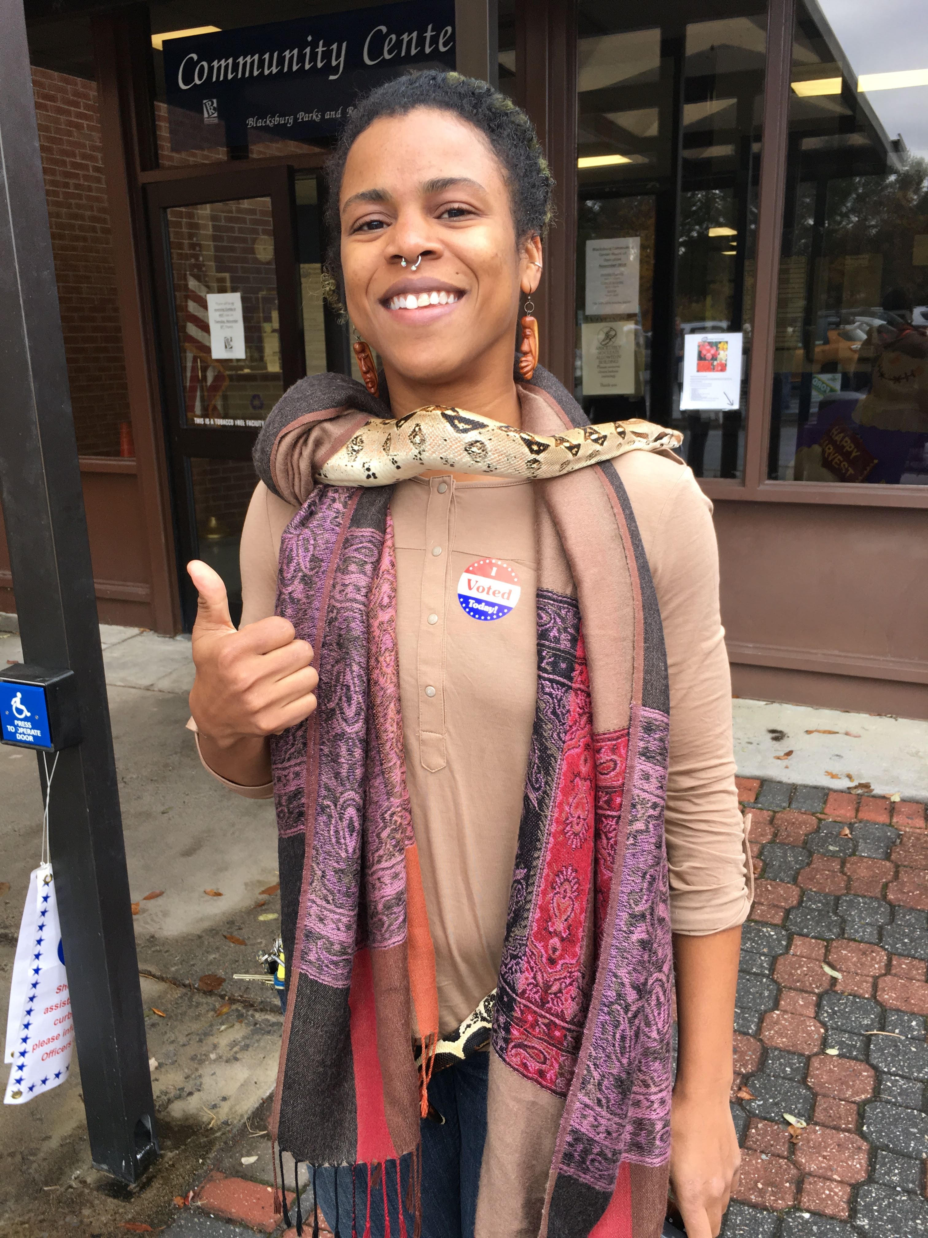 This photo shows a young woman wearing a light beige sweater, a pink and purple patterned scarf around her neck and dangling down her front, and a large snake around her neck. She has an "I voted" sticker on her sweater and is smiling at the camera and giving it a thumbs up.