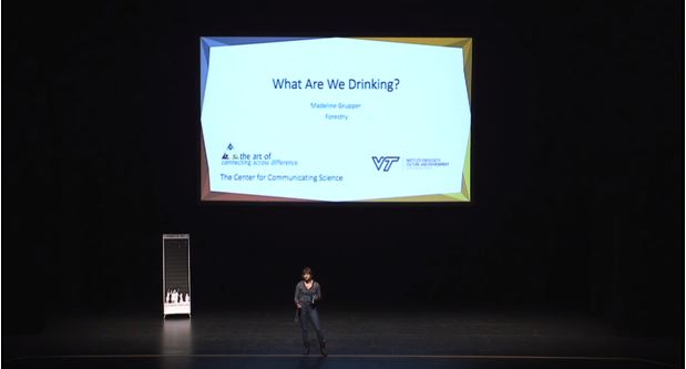 This photo shows a small figure on a large stage. in the background is a large slide reading "What Are We Drinking?" Madeline Grupper, Forestry, the art of connecting across difference, The Center for Communicating Science, VT Institute for Society, Culture and Environment.
