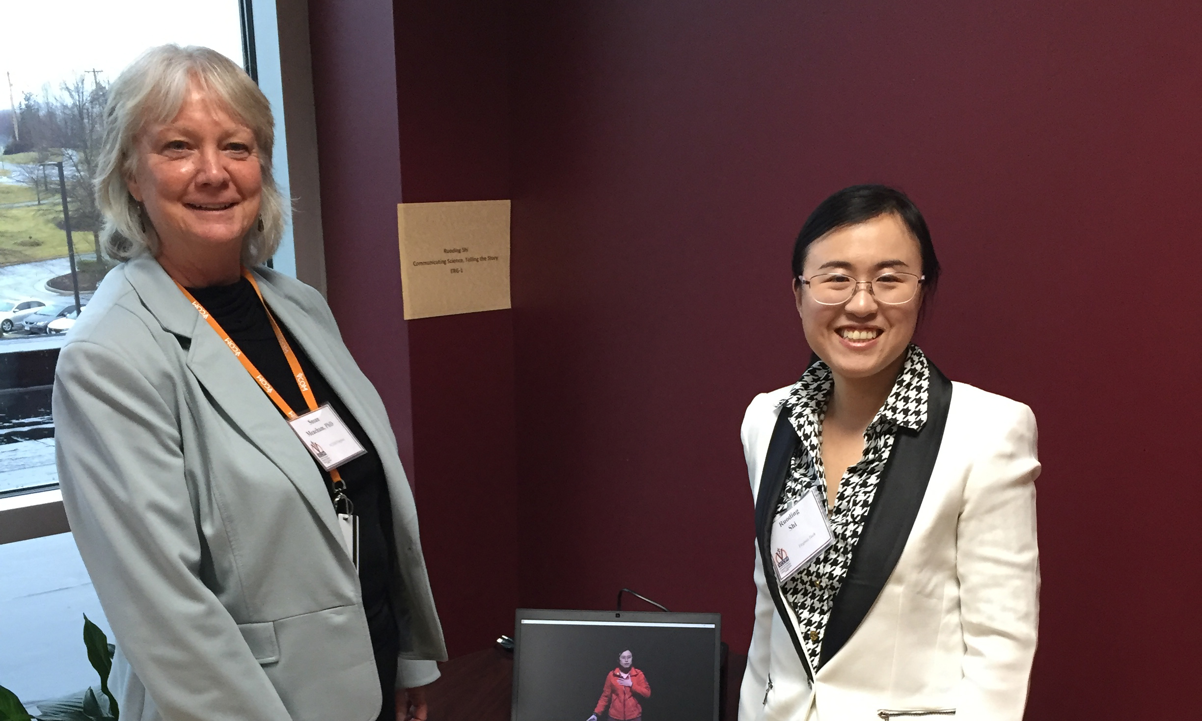 This photo shows two women standing in front of a dark maroon wall and on either side of a laptop screen. On the left is a a white woman with gray hair. On the right is a younger Asian woman with black hair. Both are professionally dressed, smiling, and wearing name tags.