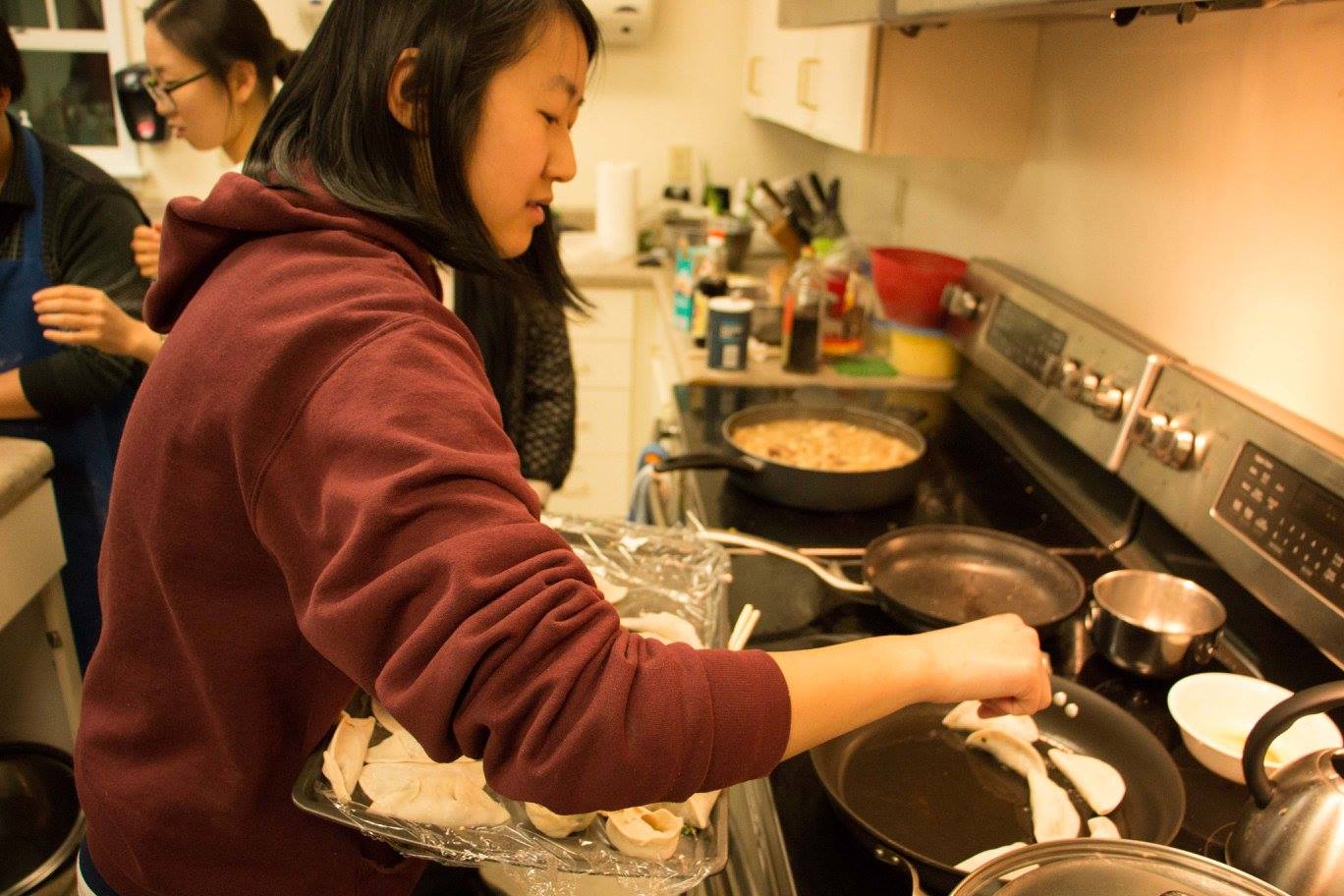 This photo shows a young dark haired woman holding a large tray of uncooked dumplings and placing a dumpling into a frying pan on a stovetop in a kitchen.