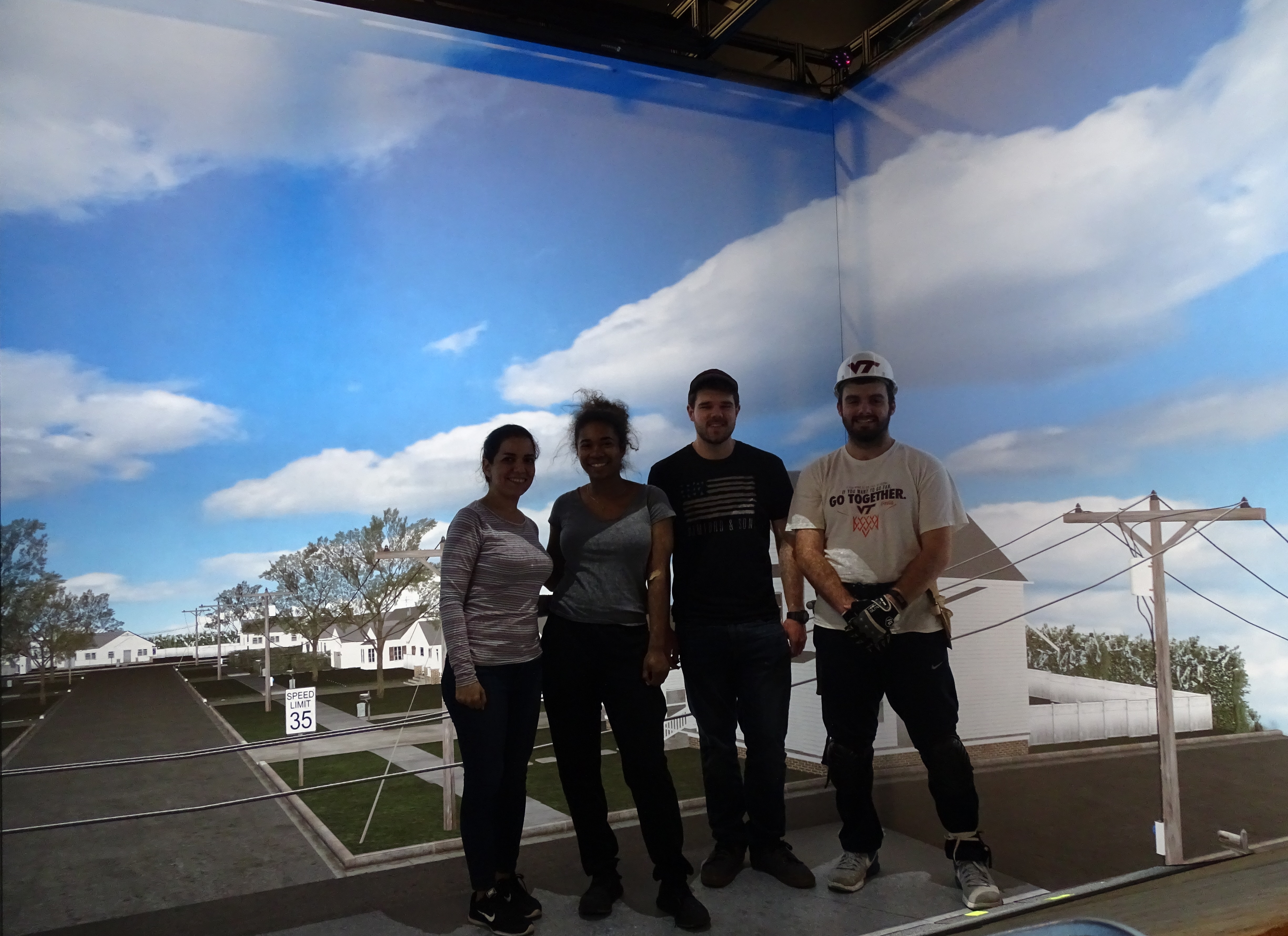 This photo shows two young women and two young men in a virtual reality environment showing blue sky, clouds, electric lines, houses, and trees.