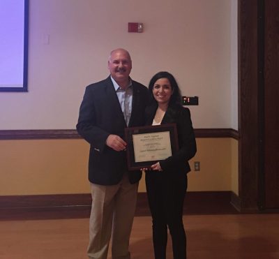 This photo shows a middle aged balding man with a young dark haired woman holding a framed award certificate. The setting is indoors. Both are dressed in business attire.