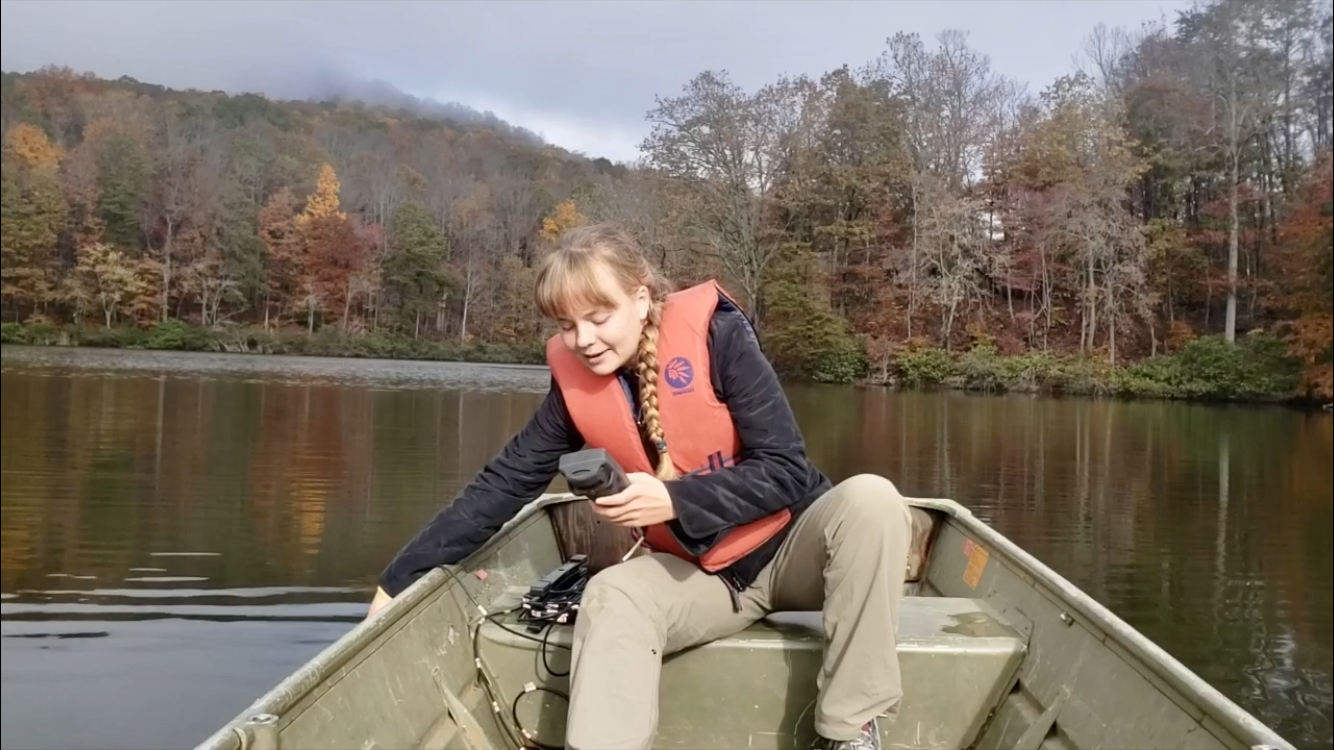 This photo shows a young Caucasian woman with a blond braid and bangs wearing beige pants, a dark jacket, and an orange life jacket. She is sitting on the back seat of a rowboat on a calm expanse of water with trees in autumn foliage behind her.