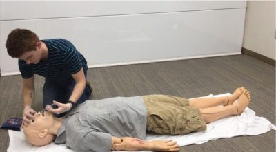 This photo shows a life-like male manikin dressed in khaki shorts and a gray t-shirt and lying on a white sheet on a carpeted floor. Kneeling at the manikin's head is a young white male dressed in a blue-and-black striped t-shirt and dark pants. He has what appears to be a syringe in his hand and is inserting it into the manikin's nostrils while using the other hand to tilt back the manikin's head and steady it.