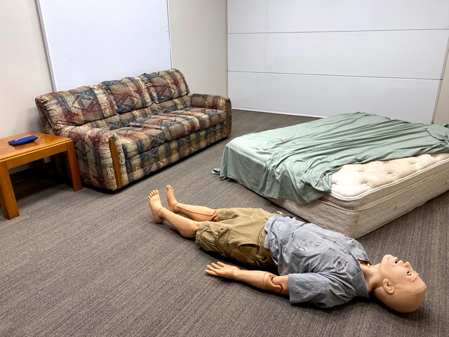 This photo shows a life-like male manikin dressed in khaki shorts and a gray t-shirt and lying on a carpeted floor. Also in the room are a sofa, an end table, and a mattress with a green sheet partially removed.