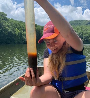 This photo shows a young Caucasian woman with blond hair dressed in shorts, a black t-shirt, a blue life vest, and an orange and maroon Virginia Tech Hokies ball cap. She is seated in a boat and holding a tall plastic tube filled with water and sediment. Behind the body of water in which the boat sits are green trees and blue sky.