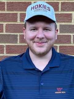 This photo shows the head and shoulders of a young white male wearing a Hokies/Virginia Tech ball cap and a dark blue polo shirt. His bearded face is smiling.