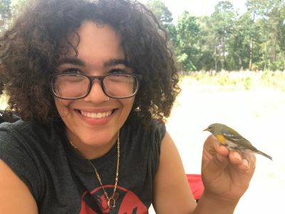 This photo shows a young woman with light brown skin and black hair, glasses, and a black t-shirt.  She is holding a small olive-gray-and-gold bird in one hand and smiling at the camera.
