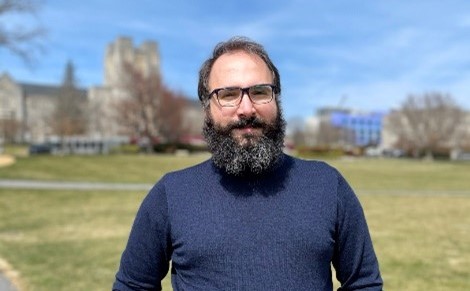 This photo shows a young man with dark hair and a full beard dressed in a dark blue long-sleeved shirt. He is smiling at the camera and behind him are campus buildings.