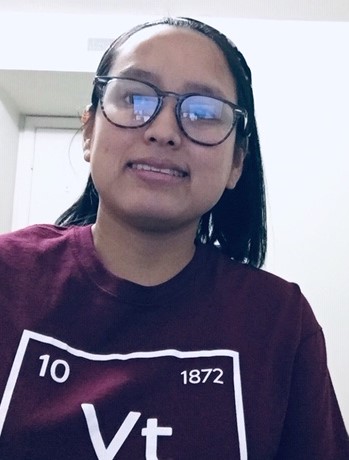 This photo shows a young woman with light brown skin, black hair, glasses, and a maroon t-shirt that is made to look like one of the elements on the periodic table. The "element" is "Vt," with numbers 10 and 1872 associated with it. She is smiling at the camera and only her head and shoulders are in the photo.