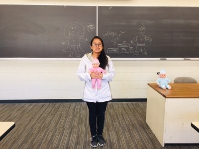 This photo shows a young woman with light brown skin, black hair, glasses, a white jacket, and black pants holding a baby doll dressed in pink. She is standing in front of a chalkboard in a classroom. On a desk sits another baby doll, this one dressed in blue.