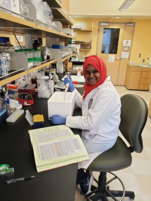 This photo shows a young woman with brown skin wearing a red head covering, a white lab coat, and blue lab gloves seated at a bench in a laboratory.