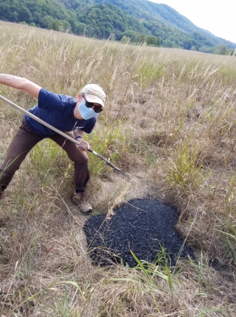 This photo shows a young light-skinned womandressed in brown pants, a navy blue t-shirt, a blue face mask, and a beige ballcap. She is standing in a meadow and poised to poke or shovel or attack a pile of black charcoal. In the background are green mountains. 