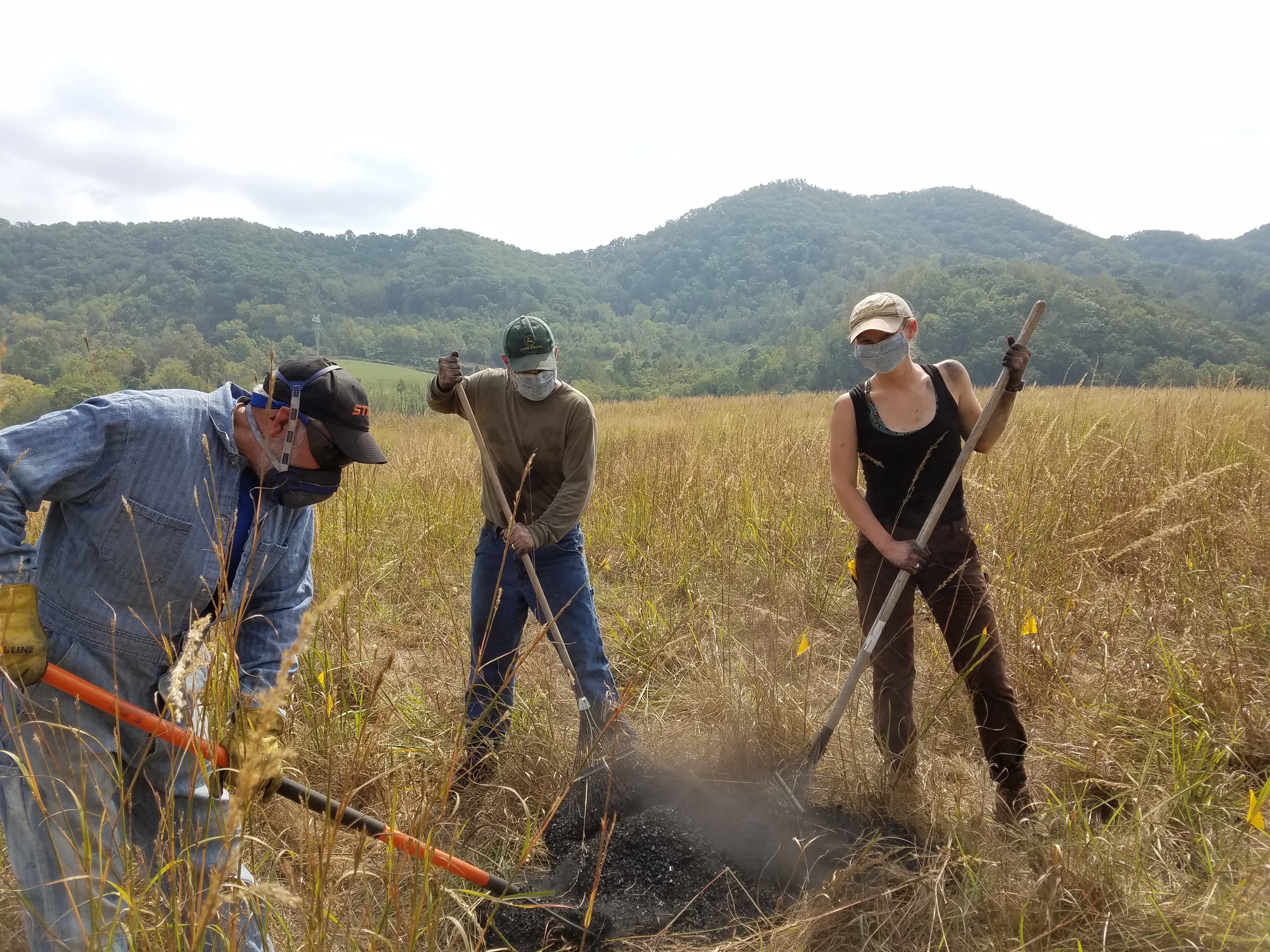 This photo shows two men and a woman, all light-skinned and clad in ball caps, shirts, pants, and face masks, shoveling or raking a pile of black charcoal in the middle of a meadow with green tree-covered mountains in the background.