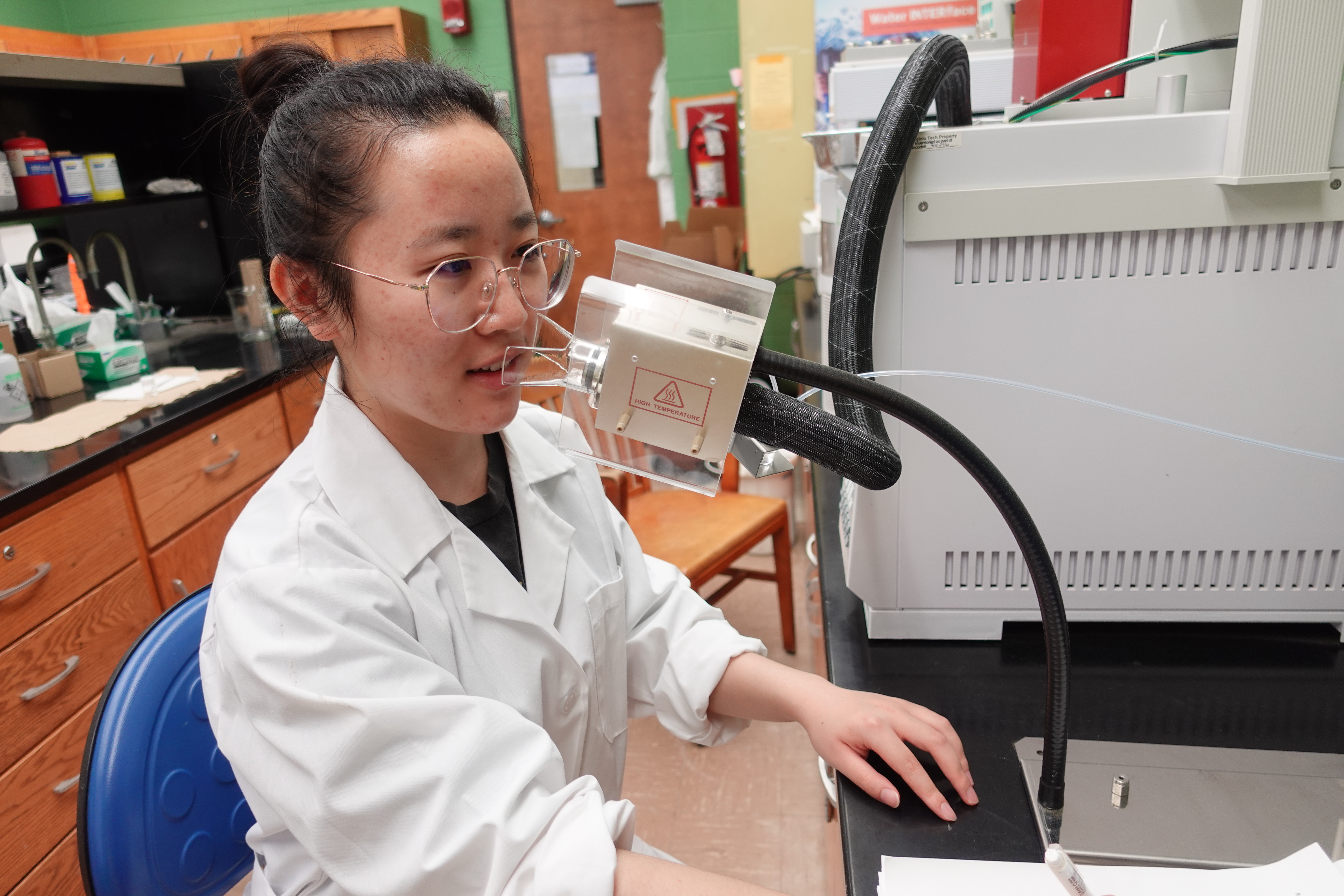 This photo shows a young Asian woman with her hair up in a bun and wearing a white lab coat. She is in a laboratory and has her fae close to a device that allows her to sniff and smell samples.. 