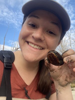 This photo shows a smiling young white woman wearing a baseball cap and an orange v-neck t-shirt. She is holding in one muddy hand a small turtle.