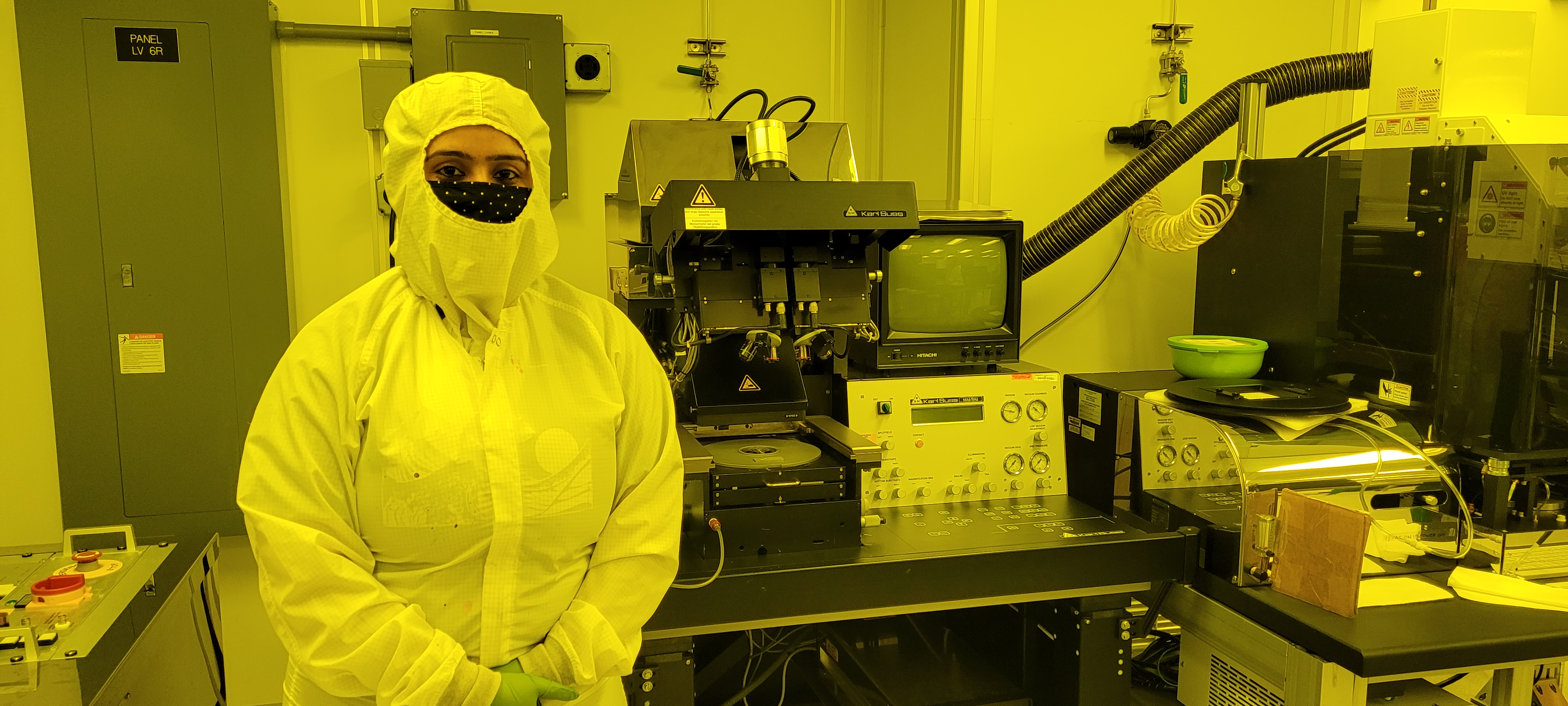 This photo shows a woman from the waist up clad in a white lab garment that covers all but her nose and eyes. She is also wearing a black face mask, so only her eyes, eyebrows, and a bit of forehead are visible. She is in a laboratory with screens, tubing, and equipment in the background of the photo.