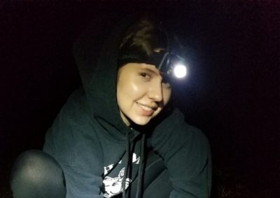 This photo shows the upper torso and head of a young white woman wearing a black hooded sweatshirt and an illuminated headlamp. She is smiling at the camera.