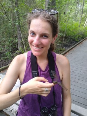 This photo should a young white woman smiling at the camera. A drogonfly is perched on one finger. She is wearing binoculars around her neck, glasses on top of her head, and a purple tank top.