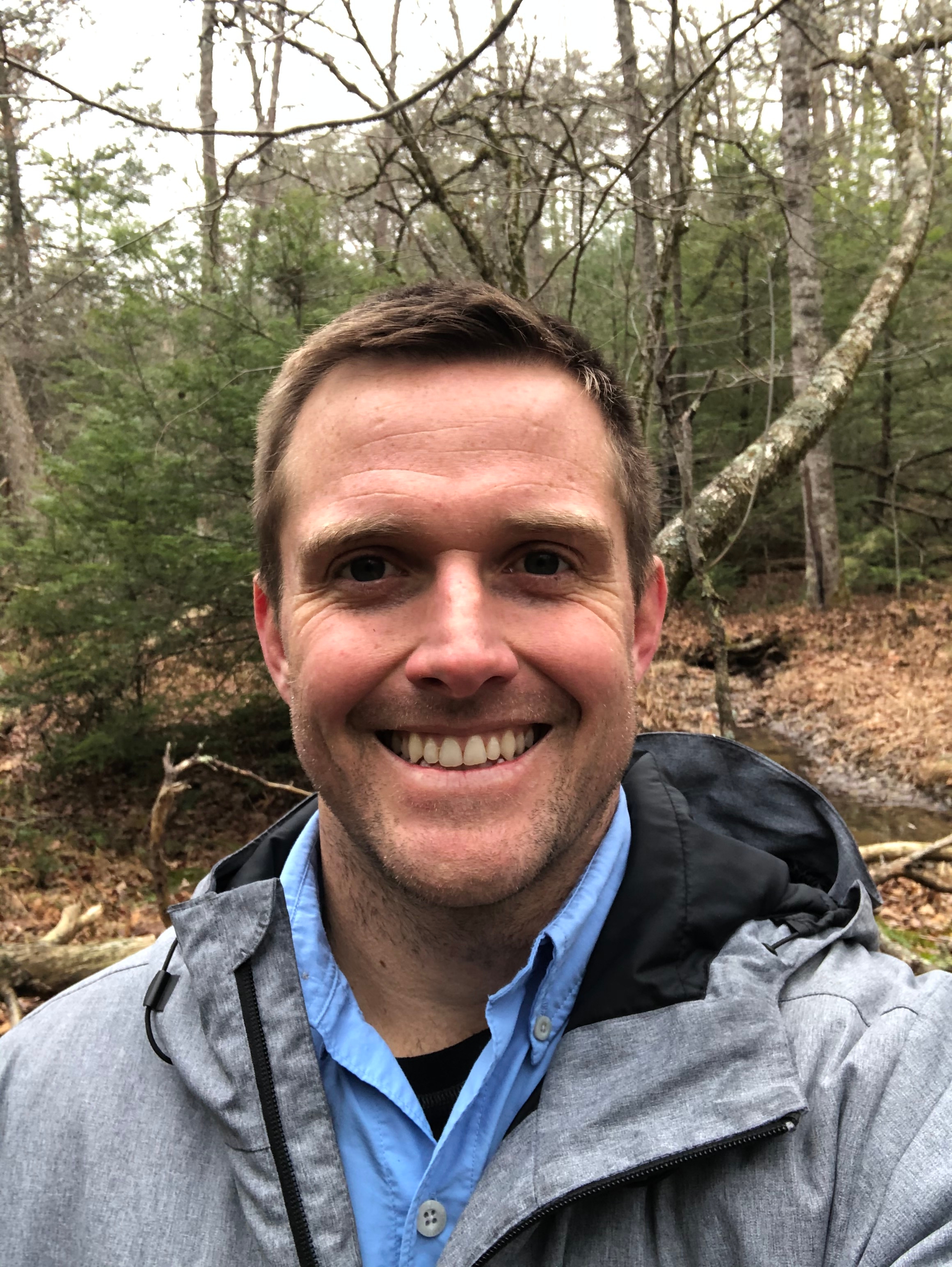 This photo shows a young white male dressed in a blue shirt with a gray hooded windbreaker over it. He is smiling at the camera. The background is scrub forest.