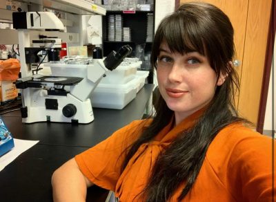 This photo shows a young white woman with long dark brown hair wearing an orange short-sleeved top and smiling at the camera. She is seated at a black lab bench and a microscope and other lab equipment is visible in the background.