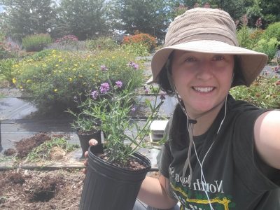 This photo shows a young white woman in a black t-shirt and beige sun hat holding a black plastic pot containing a purple-blooming plant in it. She is smiling at the camera and behind her is a large garden with many blooming plants.