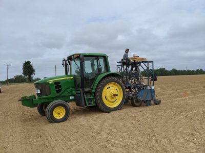 This photo shows a John Deere green and yellow tracker with another contraption attached and a person sitting on top of it. It is all sitting on a large field of dirt.