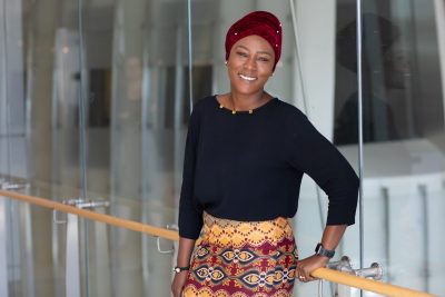 This photo shows a young Black woman with a red turban, a black top, and colorful print skirt of yellows, blues, and reds, smiling at the camera and leaning against a railing attached to glass windows or walls. 