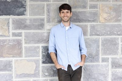 This photo shows a young white male with dark hair and a scanty beard. He wears a blue shirt, sleeves rolled up and hands in pants pockets, and is smiling at the camera.