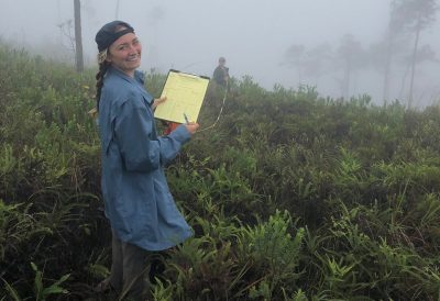 This photo shows a young white woman with blond braids standing in a field of thigh-high plants. She is holding a yellow notepad. Dense fog surrounds her, and another person can be seen dimly in the background.